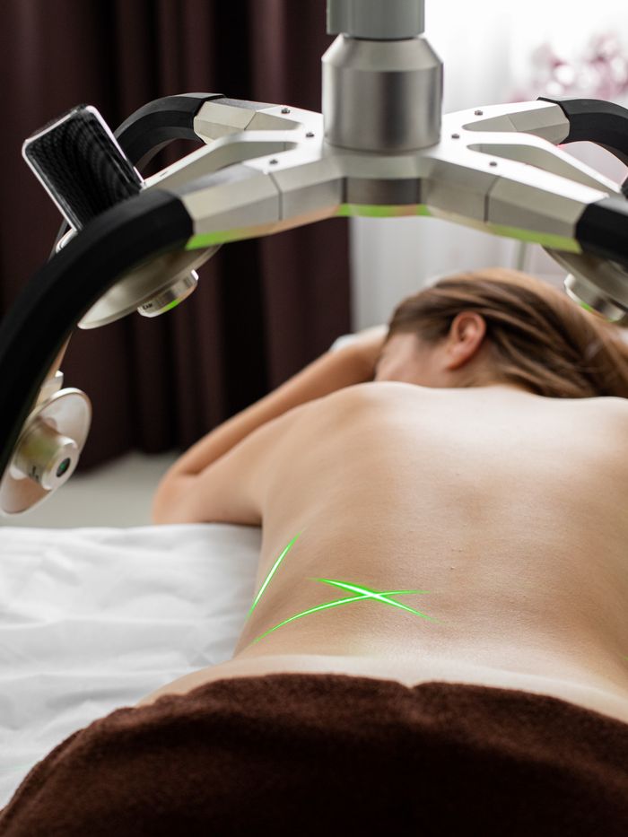 Back view of anonymous topless female client lying on medical table under laser fat loss machine during liposuction treatment in clinic