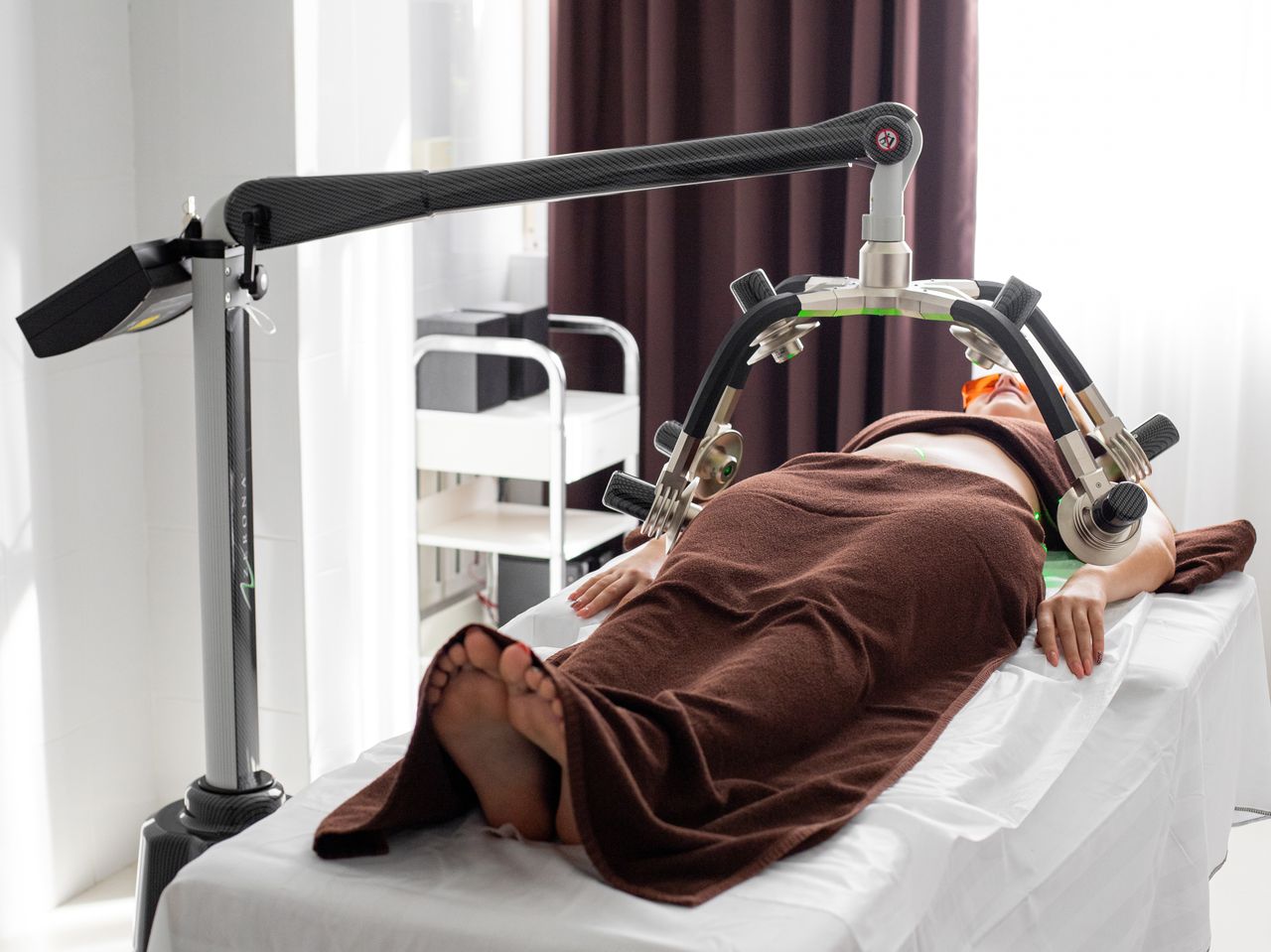 Faceless female patient covered with towels lying under laser fat loss machine during treatment in light medical clinic with curtains on window