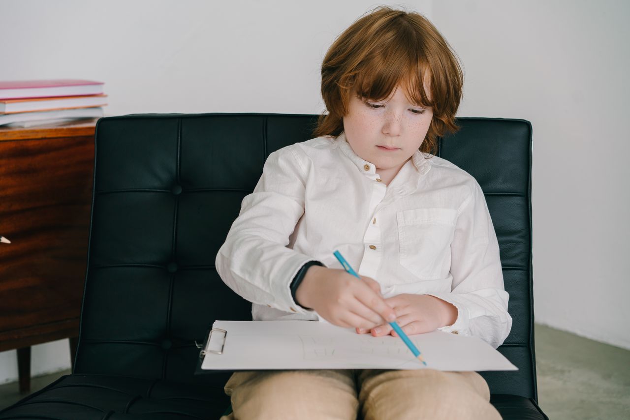 Services Child with red hair concentrates on drawing with colored pencils while sitting on a black chair.